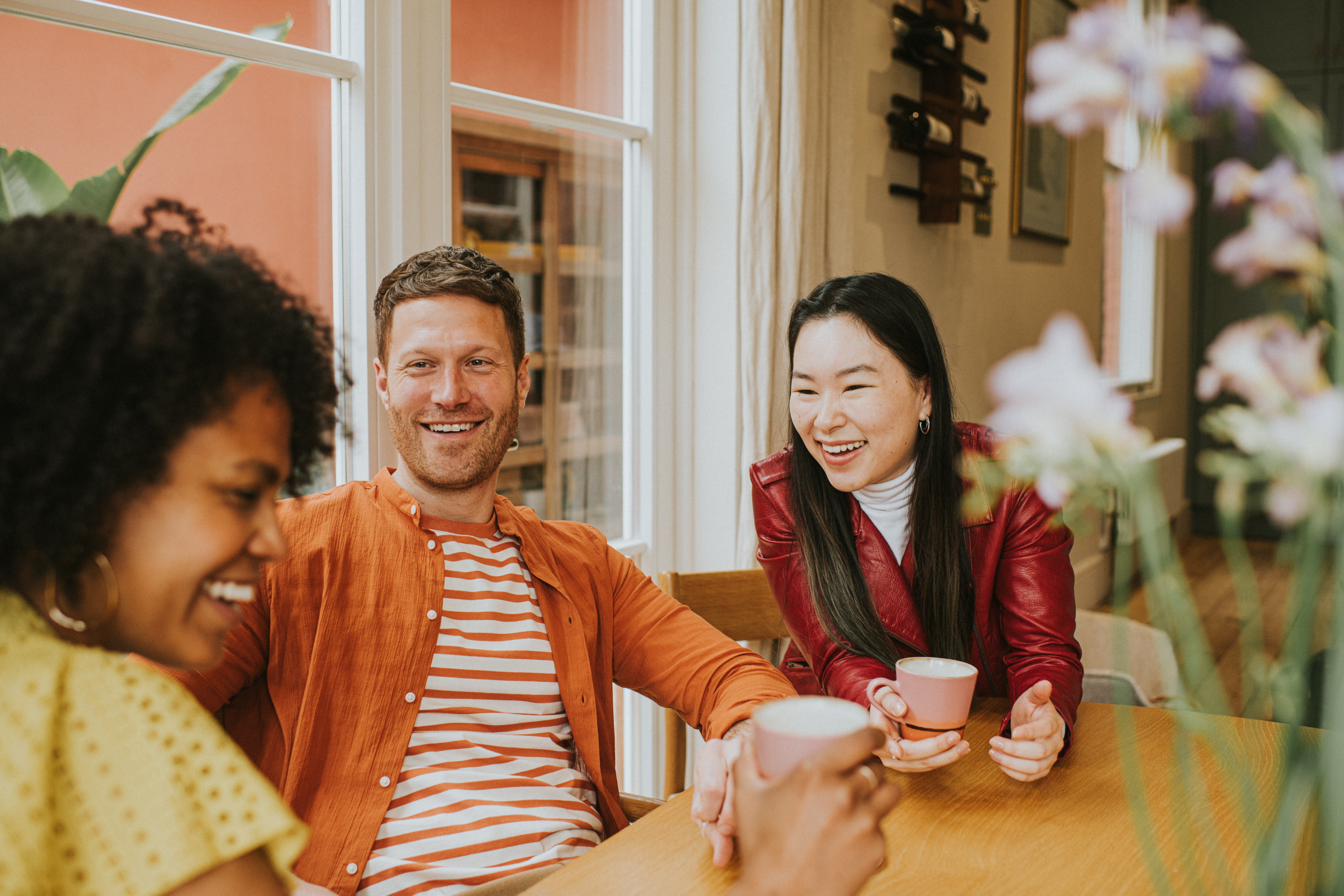 Three young people sit around a table and giggle as they have a lighthearted discussion and drink coffee / tea Programs & Services